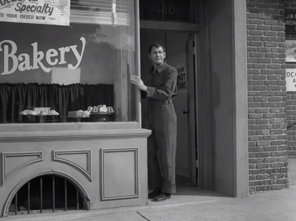 B&W still of Twilight zone episode, with a man standing in the doorway to a city scape bakery