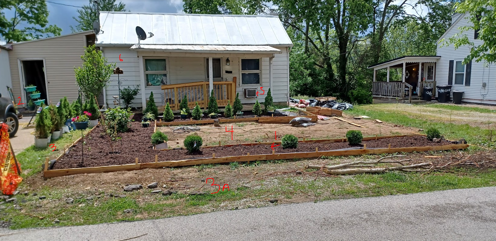 House with horseshoe raised beds and center box of dirt. Swarf spruce tree row in back, blackberry bushes to the left and 5 center hedges near the road.