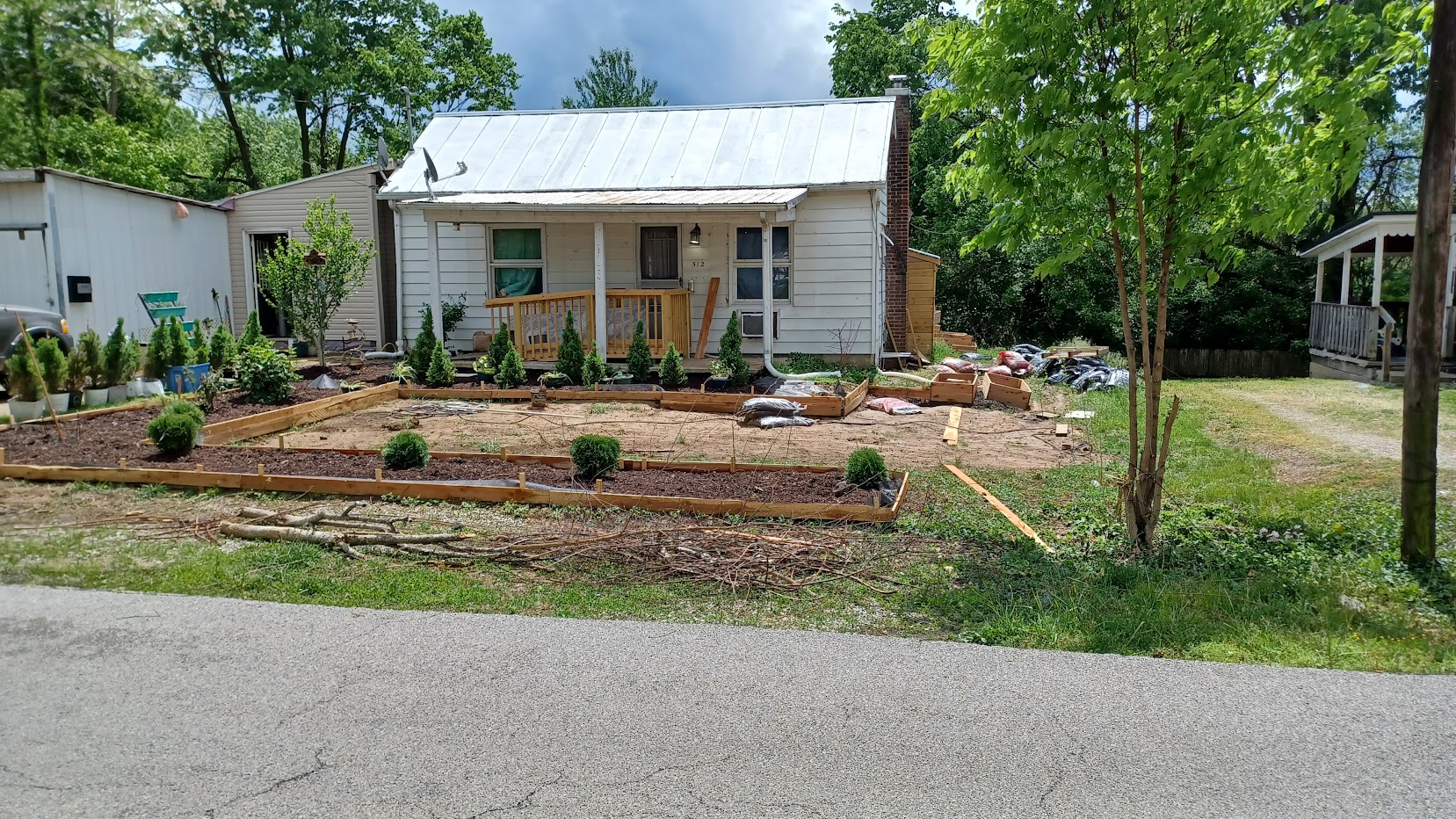 House with horseshoe raised beds and center box of dirt. Swarf spruce tree row in back, blackberry bushes to the left and 5 center hedges near the road.