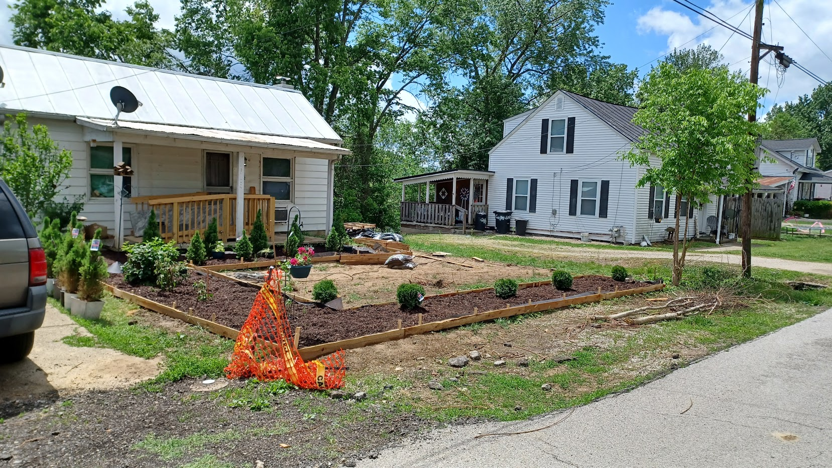 House with horseshoe raised beds and center box of dirt. Swarf spruce tree row in back, blackberry bushes to the left and 5 center hedges near the road.