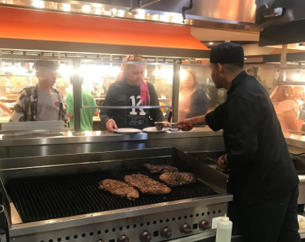 Man cooking steaks on an open flame grill and two people at the windows getting them